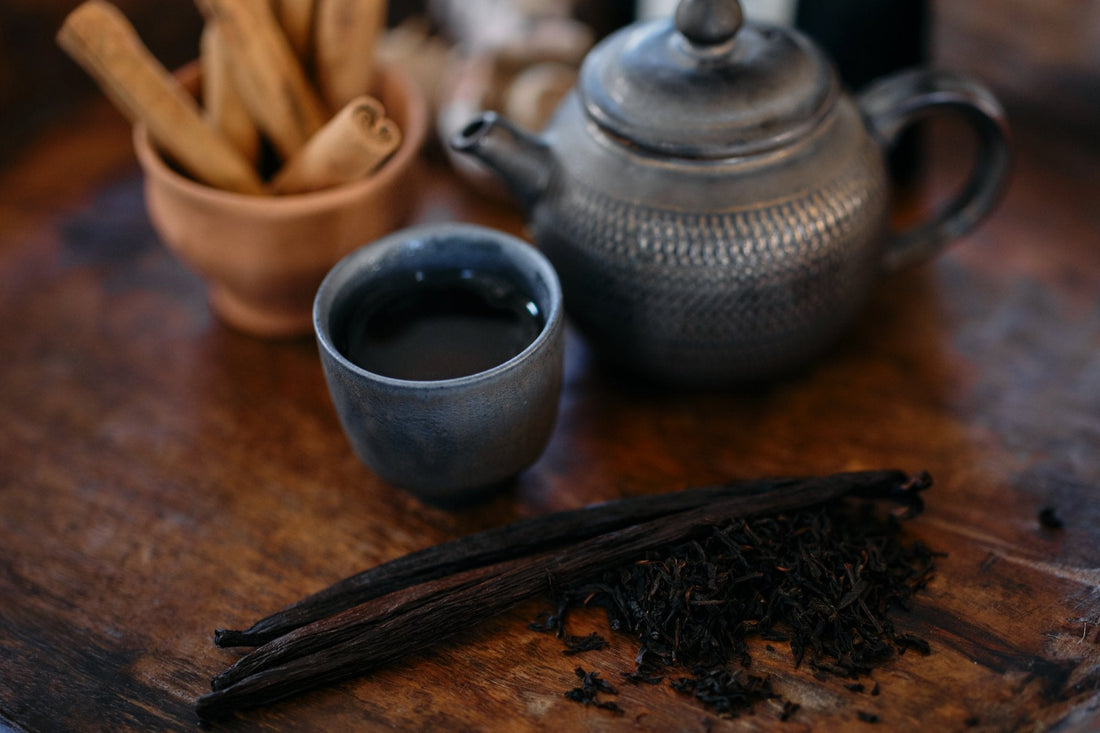 Cinnamon quills, vanilla pods and loose black tea beside a dark teapot and a small cup of tea