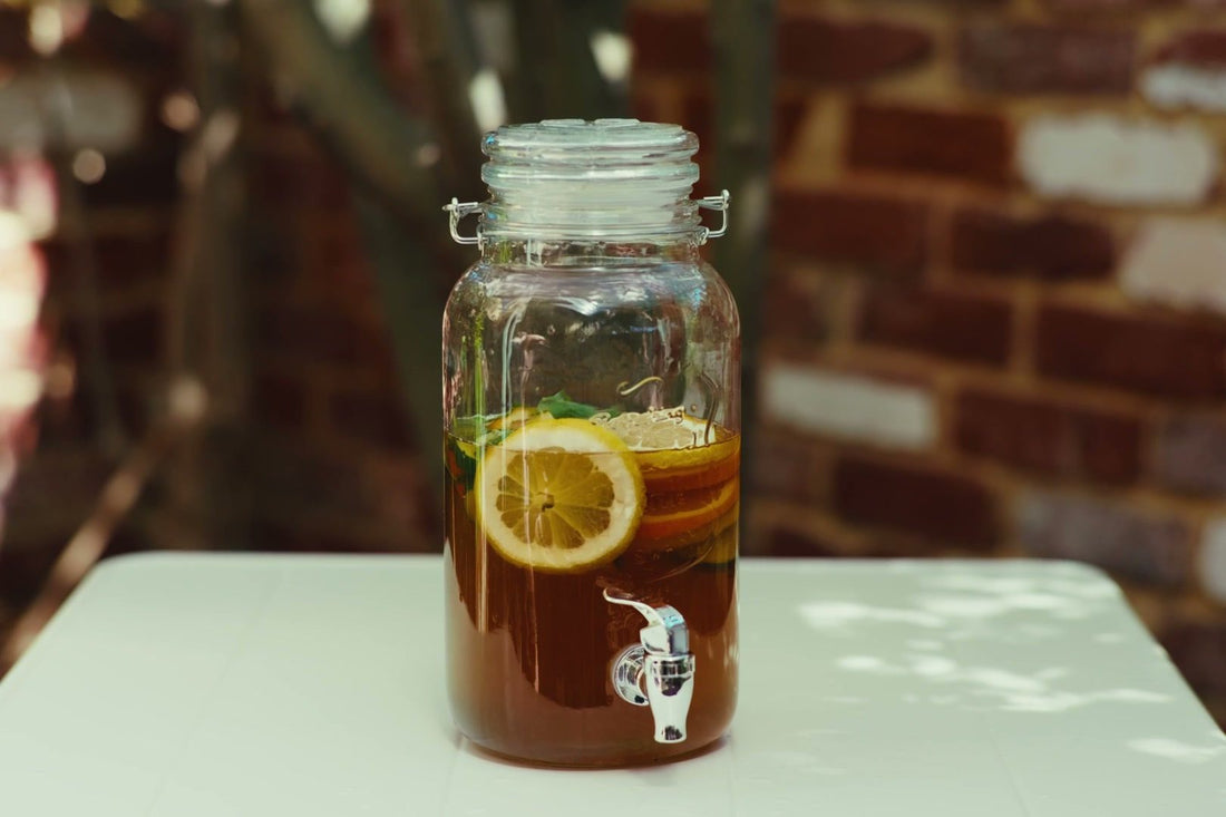 A glass drink dispenser of iced tea with a lemon slice on a sunlit table