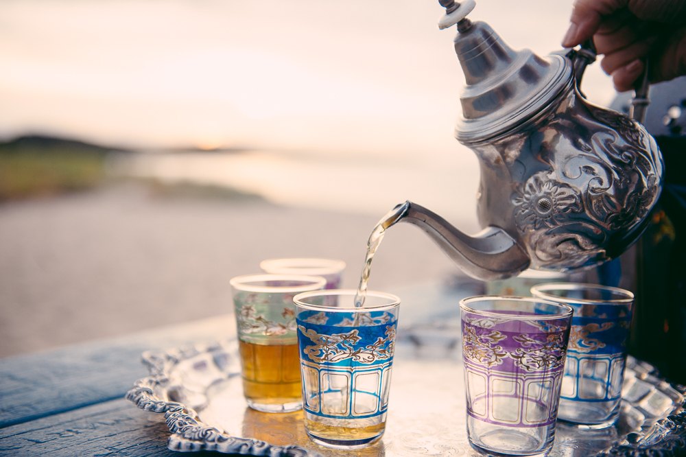 Tea poured from a silver pot into glasses on a beach table at sunset