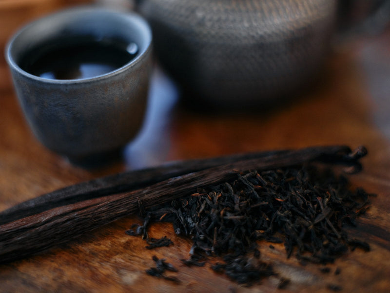 A cup of black tea beside dark loose leaf and a vanilla pod on dark timber