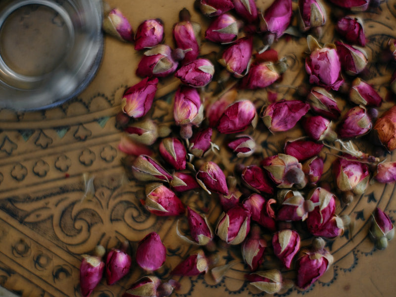 Dried rose buds spread across an engraved brass tray beside a glass