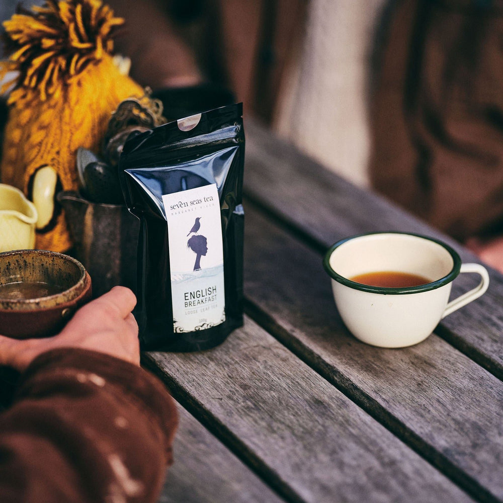 Person holding a coffee bag labeled 'English Breakfast' next to a cup of coffee on a wooden table.