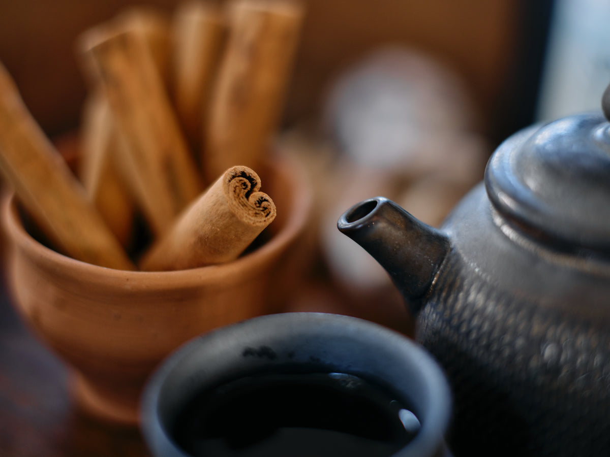 A terracotta bowl of cinnamon sticks beside a black teapot and a full cup of tea