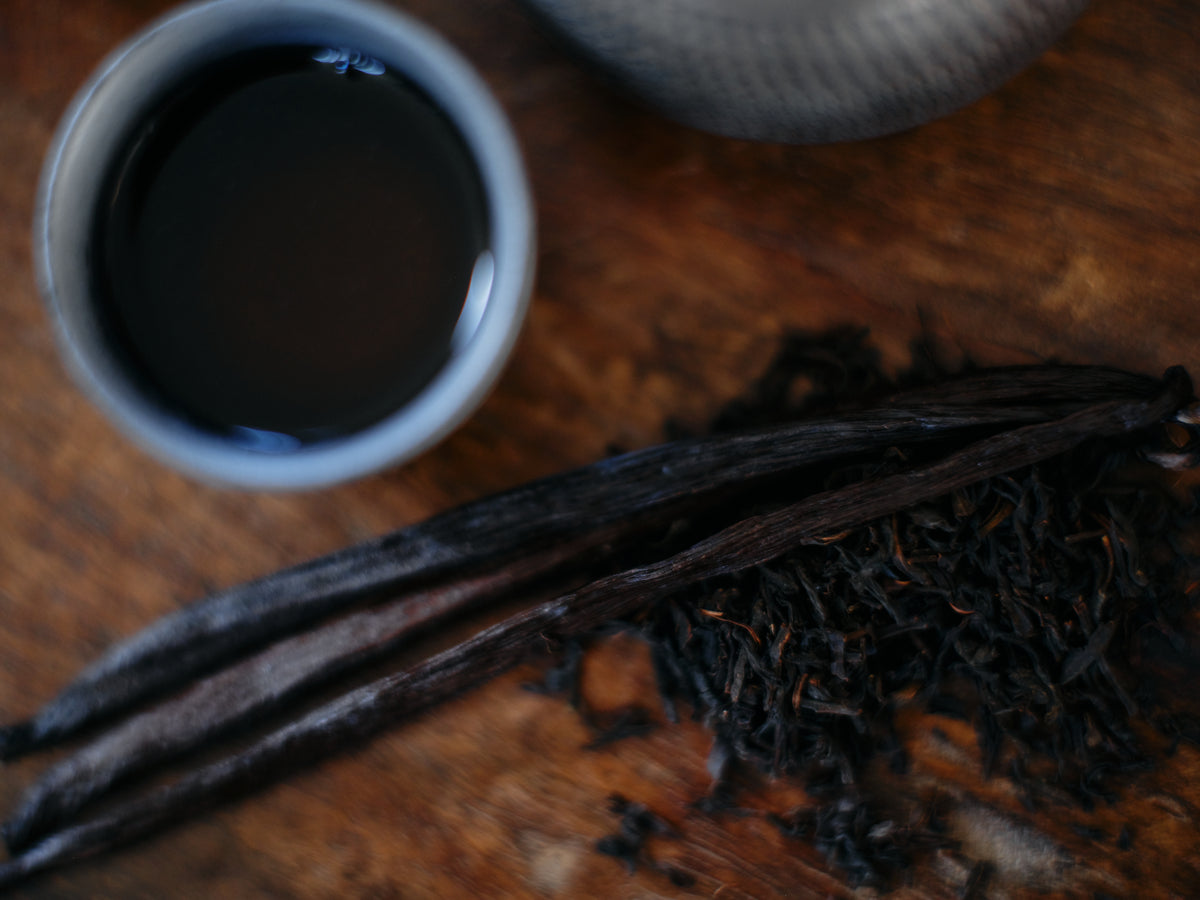 A brewed cup of black tea beside dark loose leaf and vanilla pods on a timber table